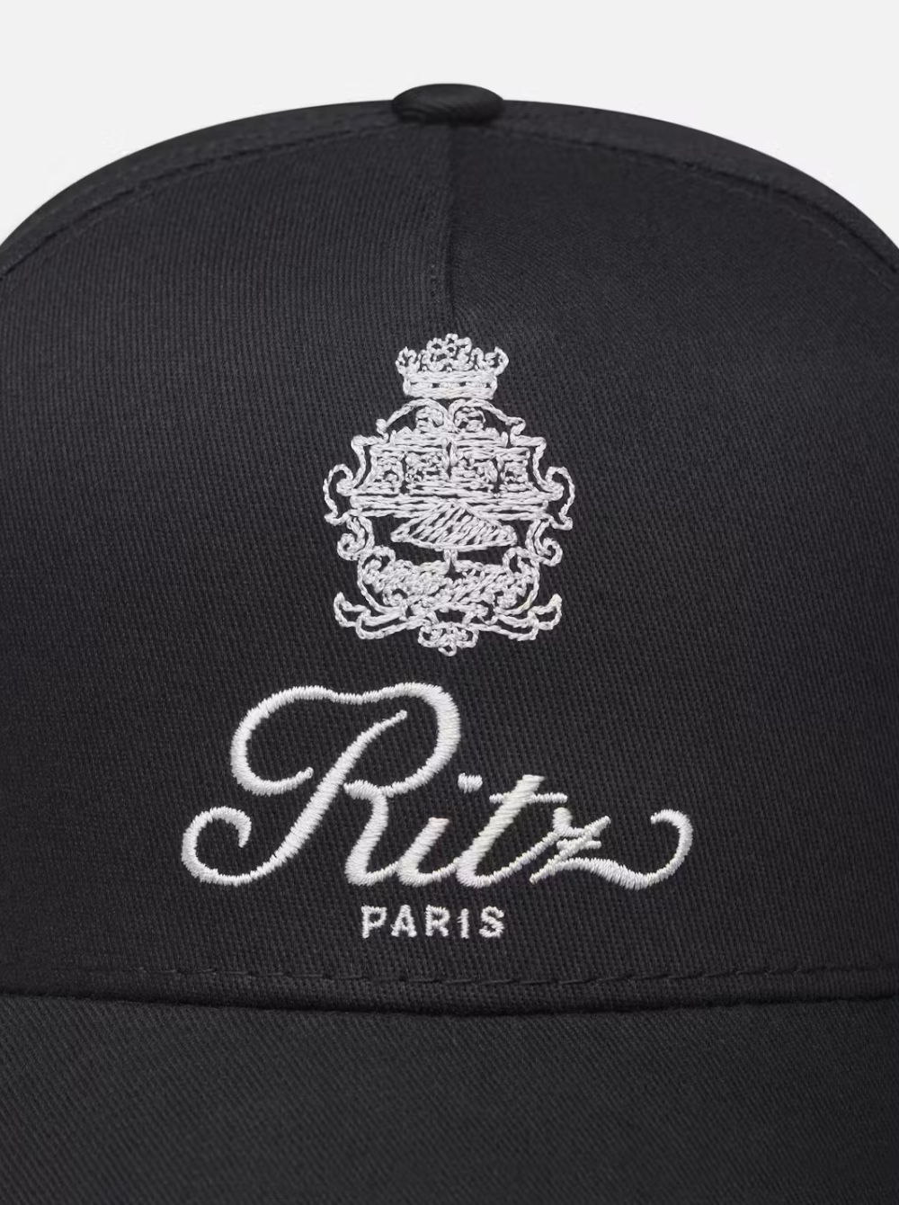 A close-up image of the Ritz Cotton Trucker, a black cotton baseball cap by FRAME, showcases an intricate white embroidered crest with the words "Ritz Paris," adding elegance and a classic touch.