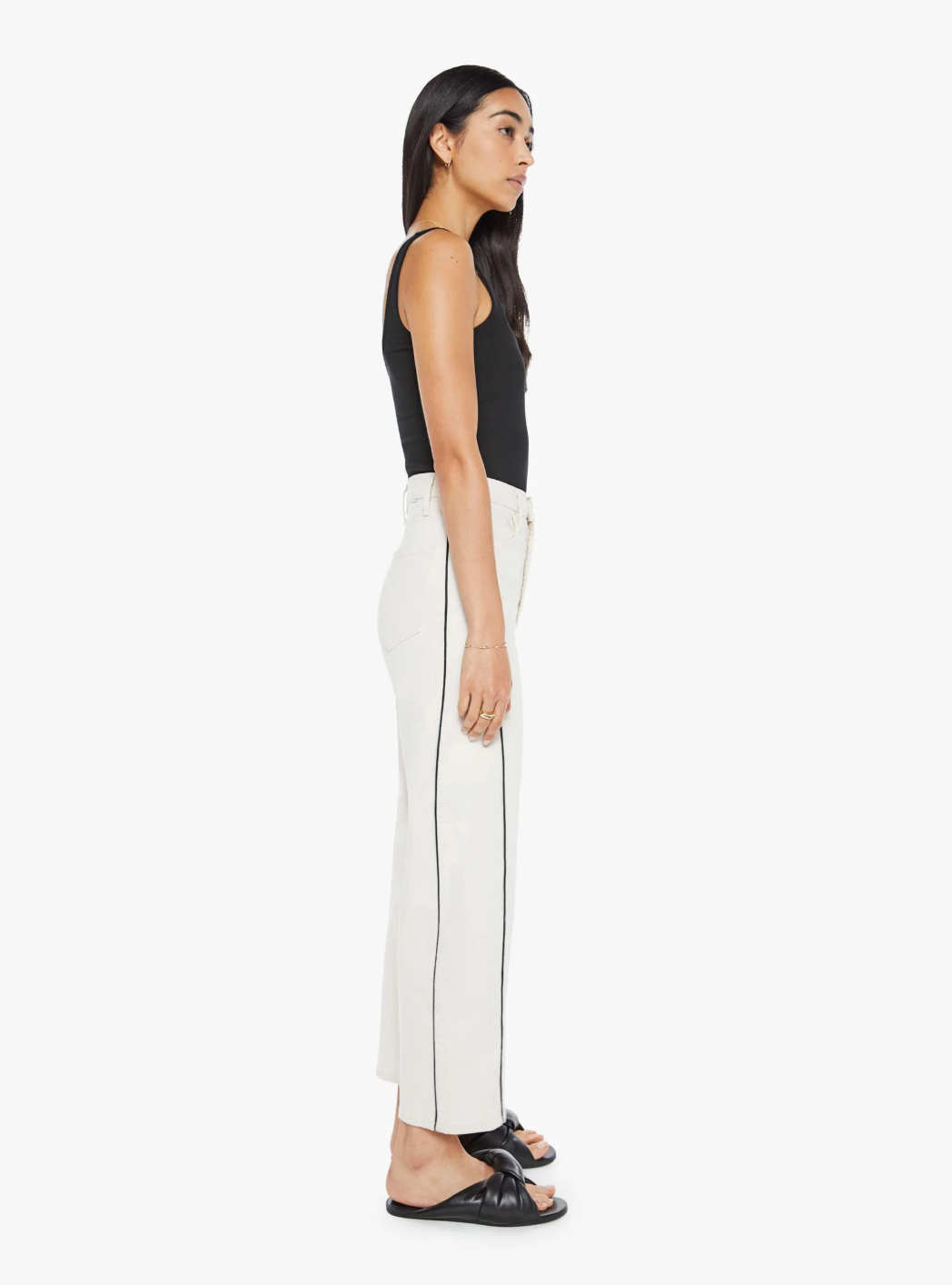 A person with long dark hair stands in profile against a plain white background, wearing a black tank top, The Half Pipe Ankle Wash: Act Natural jeans by Mother featuring vertical stripes, and black sandals.