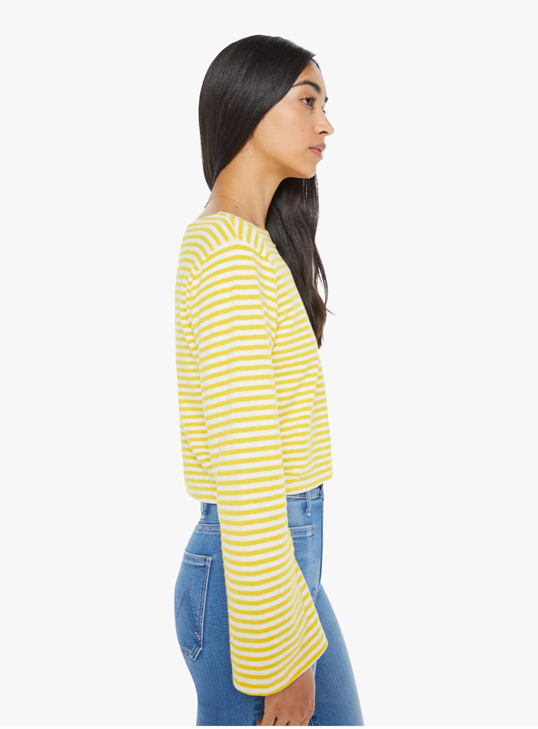 A person with long dark hair is wearing the Mother Skipper Bell LS Tee—a yellow and white striped stretch cotton boat neck top—and blue jeans. They are standing sideways facing left against a plain white background.