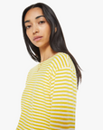 A person with long dark hair is wearing The Skipper Bell LS Tee by Mother, featuring yellow and white horizontal stripes and long sleeves, gazing at the camera with a neutral expression against a white background.