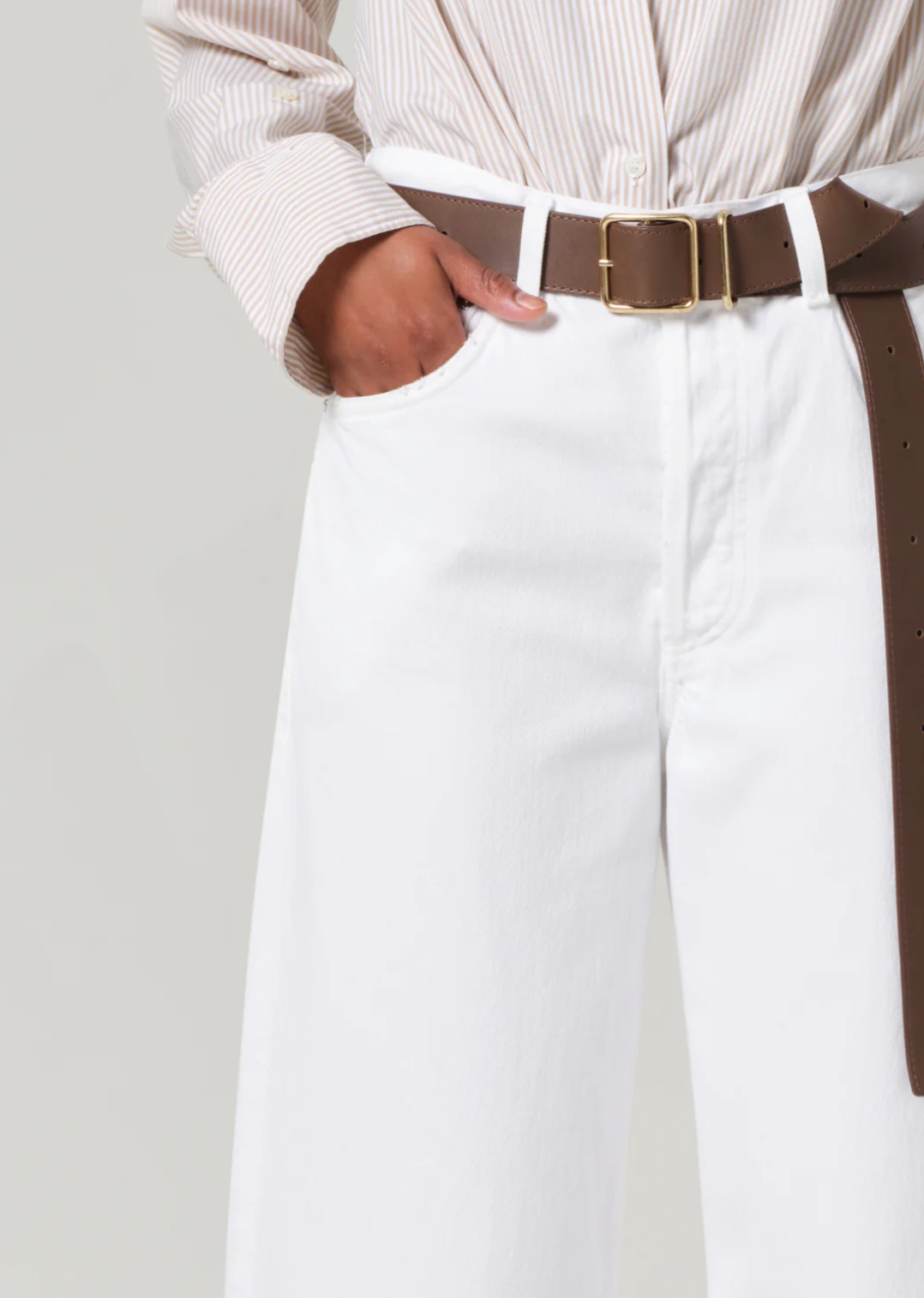 A person in a white shirt tucked into Citizens Of Humanity/AGOLDE Ayla Raw Hem Crop pants has hands in pockets. A brown leather belt with a gold buckle accents the relaxed rise of these rigid cotton trousers against a plain gray background.