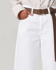 A person in a white shirt tucked into Citizens Of Humanity/AGOLDE Ayla Raw Hem Crop pants has hands in pockets. A brown leather belt with a gold buckle accents the relaxed rise of these rigid cotton trousers against a plain gray background.