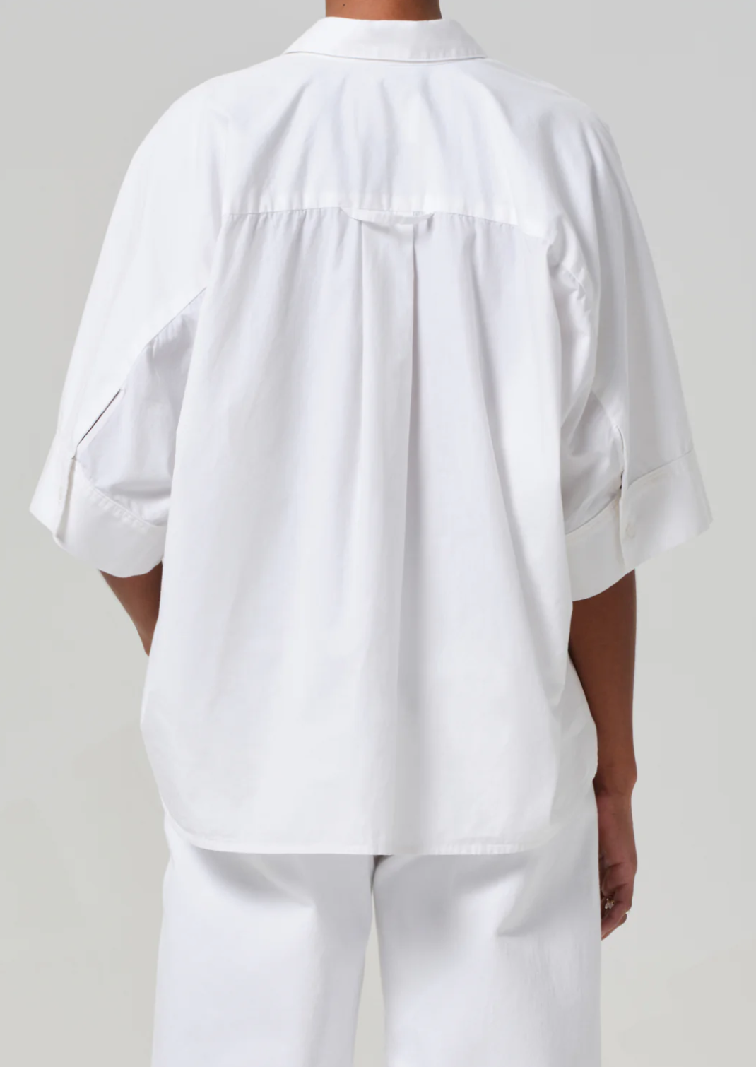 The individual is donned in a breezy cotton-poplin Claire Origami Shirt by Citizens of Humanity/AGOLDE, with oversized sleeves and a back yoke detail, paired with matching white pants. Facing away from the camera, they stand against a plain background.