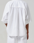 The individual is donned in a breezy cotton-poplin Claire Origami Shirt by Citizens of Humanity/AGOLDE, with oversized sleeves and a back yoke detail, paired with matching white pants. Facing away from the camera, they stand against a plain background.