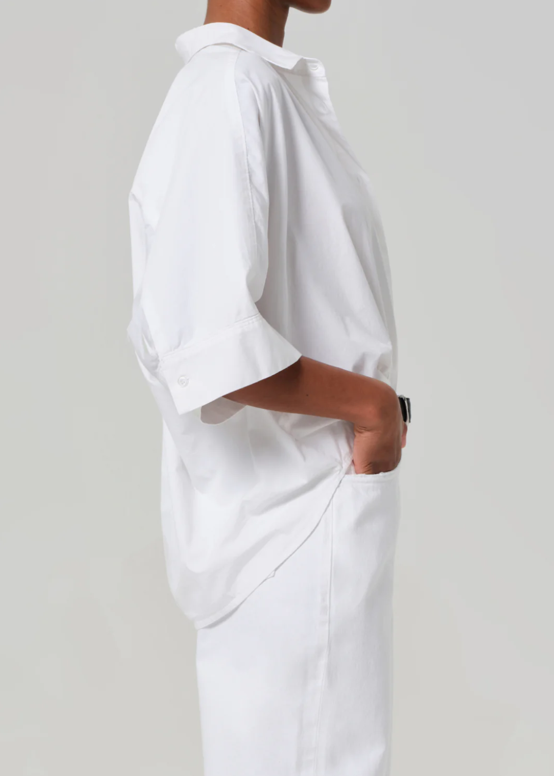 Wearing the Claire Origami Shirt by Citizens Of Humanity/AGOLDE, an individual stands in profile with hands pocketed. The classic pointed collar adds elegance to their loose-fitting white button-up and matching pants, with the cotton-poplin fabric draping effortlessly against a light gray backdrop.