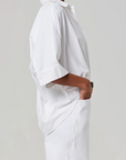 Wearing the Claire Origami Shirt by Citizens Of Humanity/AGOLDE, an individual stands in profile with hands pocketed. The classic pointed collar adds elegance to their loose-fitting white button-up and matching pants, with the cotton-poplin fabric draping effortlessly against a light gray backdrop.