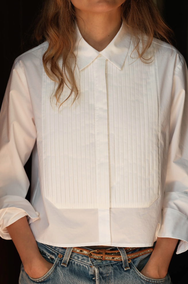 A woman is styled in Cissa's "The Tuxedo Shirt," a white tuxedo design with a pleated bib and rolled-up sleeves, paired with blue jeans. Her long hair flows freely as she stands with hands in pockets, face unseen.