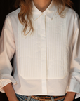 A woman is styled in Cissa's "The Tuxedo Shirt," a white tuxedo design with a pleated bib and rolled-up sleeves, paired with blue jeans. Her long hair flows freely as she stands with hands in pockets, face unseen.