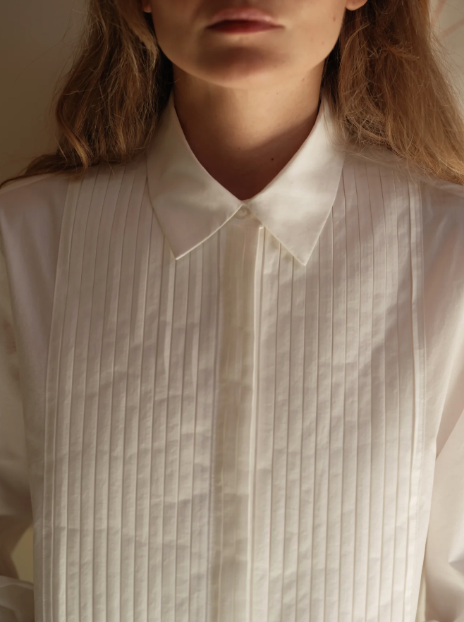 A person wears Cissa's The Tuxedo Shirt, featuring a pointed collar and pleated bib. The focus is on the upper chest and neck against a softly lit background, with long hair visible at the sides.