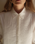 A person wears Cissa's The Tuxedo Shirt, featuring a pointed collar and pleated bib. The focus is on the upper chest and neck against a softly lit background, with long hair visible at the sides.