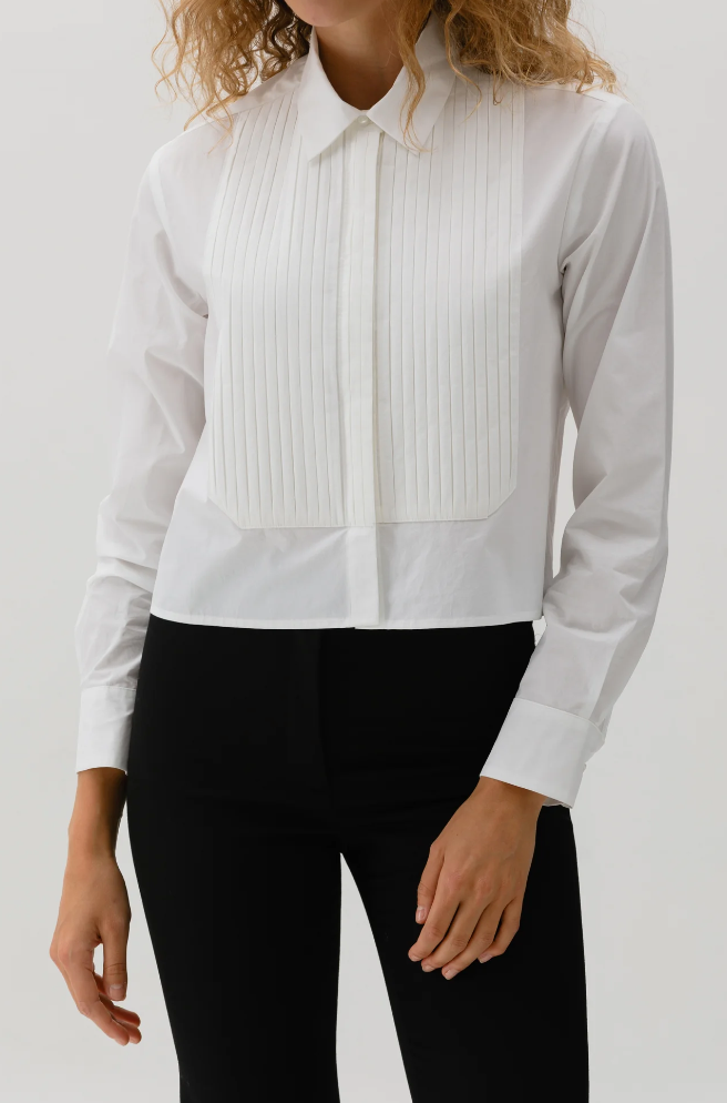 A person in The Tuxedo Shirt by Cissa, a white paper cotton shirt with a pleated bib and classic collar, pairs it elegantly with black pants against a simple backdrop, enhancing their curly hair.