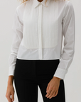 A person in The Tuxedo Shirt by Cissa, a white paper cotton shirt with a pleated bib and classic collar, pairs it elegantly with black pants against a simple backdrop, enhancing their curly hair.