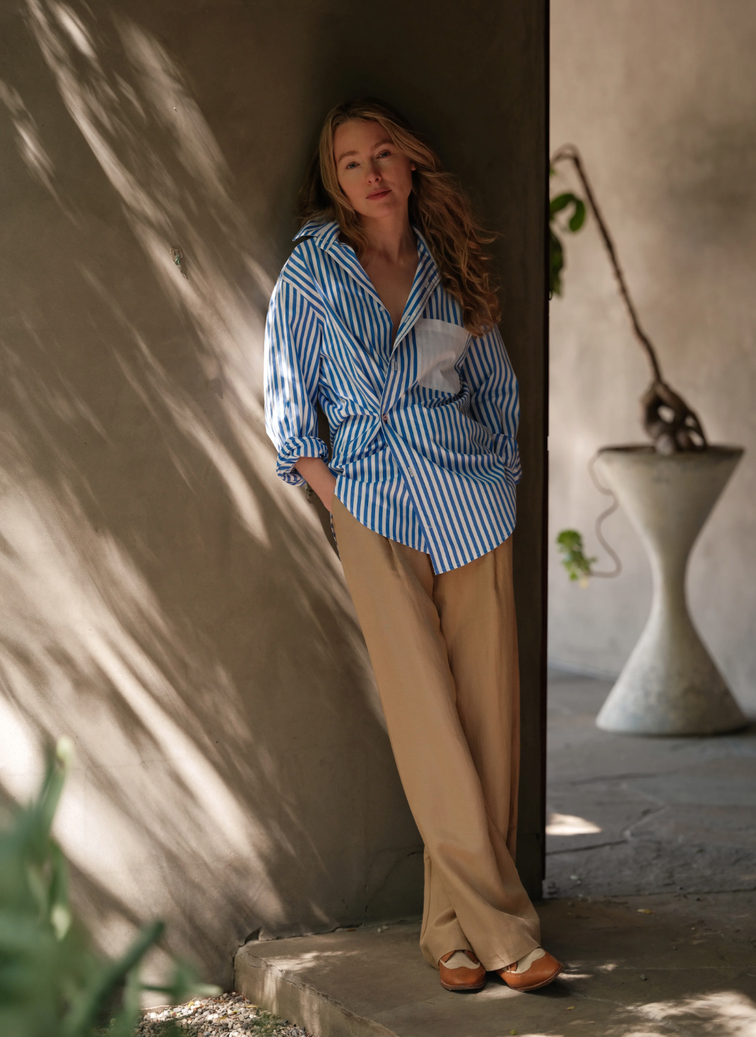 A woman with long hair leans against a wall in soft lighting, wearing The MEN'S Shirt by Cissa, featuring blue and white stripes with contrast pink buttons. Her beige pants complement the greenery as she stands near some plants and a distinctive, curved planter in the background.