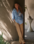 A woman with long hair leans against a wall in soft lighting, wearing The MEN'S Shirt by Cissa, featuring blue and white stripes with contrast pink buttons. Her beige pants complement the greenery as she stands near some plants and a distinctive, curved planter in the background.