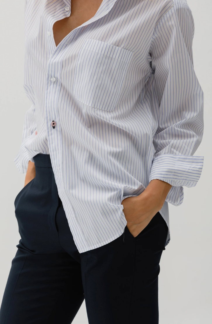 Dressed in "The MEN'S Shirt" by Cissa, featuring elegant white and blue pinstripes crafted from luxurious Giza 87 Egyptian Cotton, a person casually rolls up the sleeves and pairs it with black pants, standing with hands in pockets against a plain backdrop.