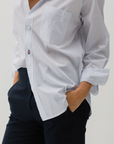 Dressed in "The MEN'S Shirt" by Cissa, featuring elegant white and blue pinstripes crafted from luxurious Giza 87 Egyptian Cotton, a person casually rolls up the sleeves and pairs it with black pants, standing with hands in pockets against a plain backdrop.