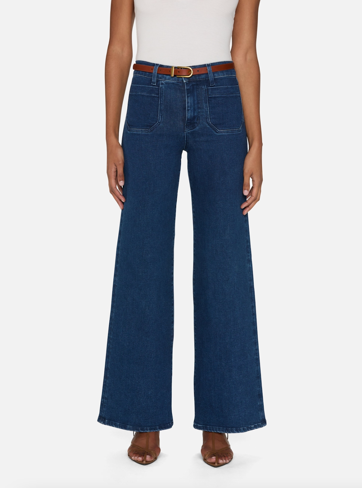 Someone is wearing FRAME&#39;s &quot;Le Slim Palazzo Modernist&quot; high-waisted, wide-leg, flare blue jeans made of super stretch denim. They have a white top tucked in with a brown belt and are standing against a plain white background in brown open-toe sandals.