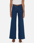 Someone is wearing FRAME's "Le Slim Palazzo Modernist" high-waisted, wide-leg, flare blue jeans made of super stretch denim. They have a white top tucked in with a brown belt and are standing against a plain white background in brown open-toe sandals.