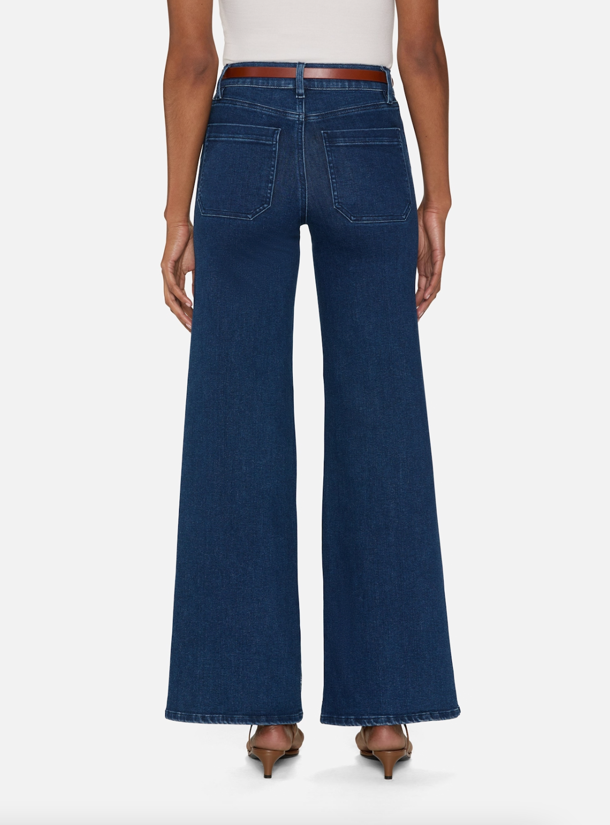 A person is seen from behind wearing FRAME&#39;s Le Slim Palazzo Modernist jeans, a high-rise wide-leg design in dark blue super stretch denim with large back pockets. They&#39;re paired with heeled shoes and a white top against a plain white background.