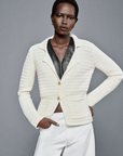 A person stands confidently with hands in pockets, wearing the FRAME Crochet Blazer over a black top and white pants. This workwear essential complements their short hair and focused expression against the plain grey background.