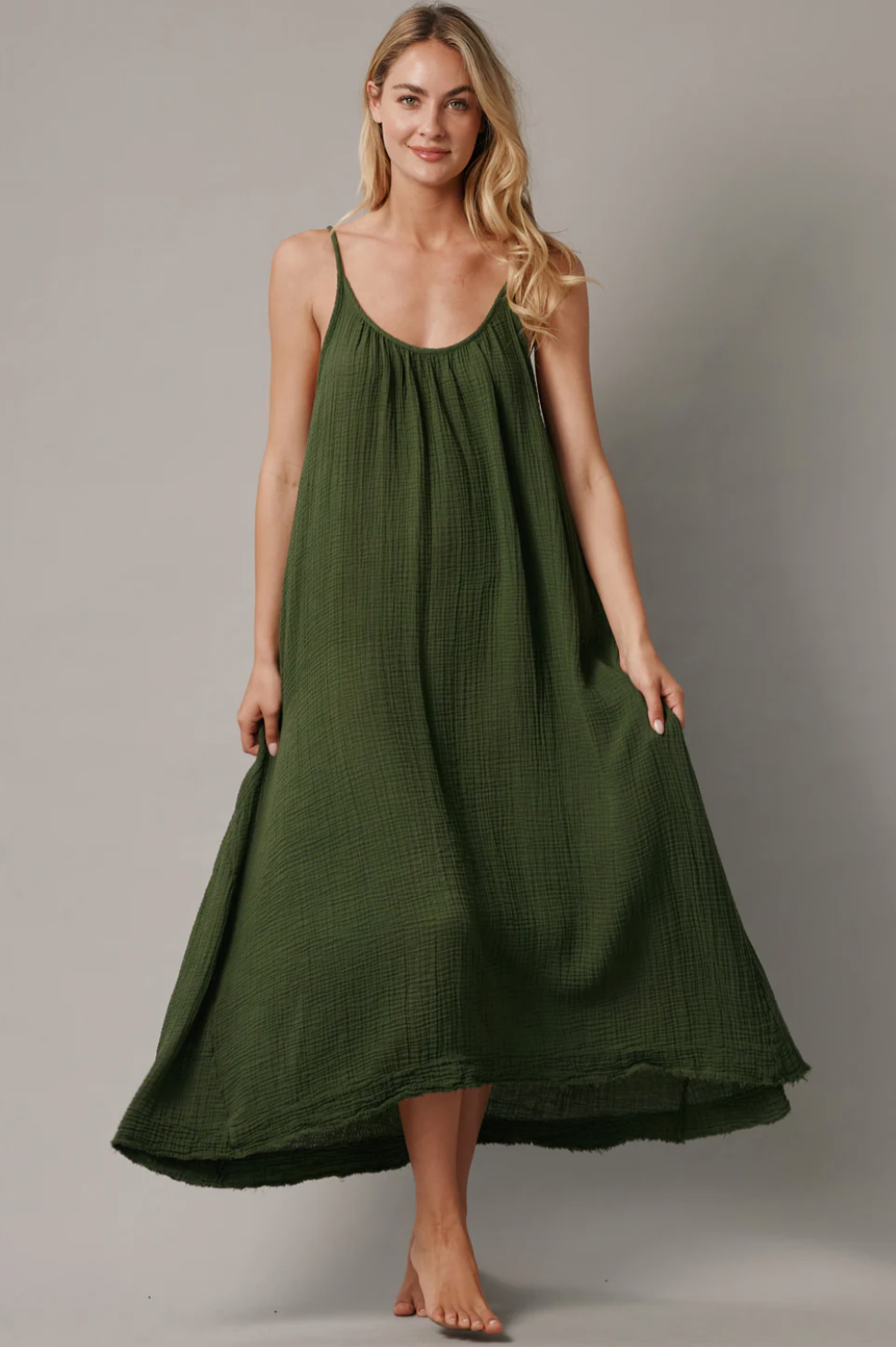 Barefoot against a plain backdrop, a woman wears the Tulum maxi by 9seed. Crafted from 100% cotton and lightweight gauze, the flowing dark green sleeveless dress elegantly drapes to ankle length as her long blonde hair cascades down.