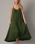 Barefoot against a plain backdrop, a woman wears the Tulum maxi by 9seed. Crafted from 100% cotton and lightweight gauze, the flowing dark green sleeveless dress elegantly drapes to ankle length as her long blonde hair cascades down.