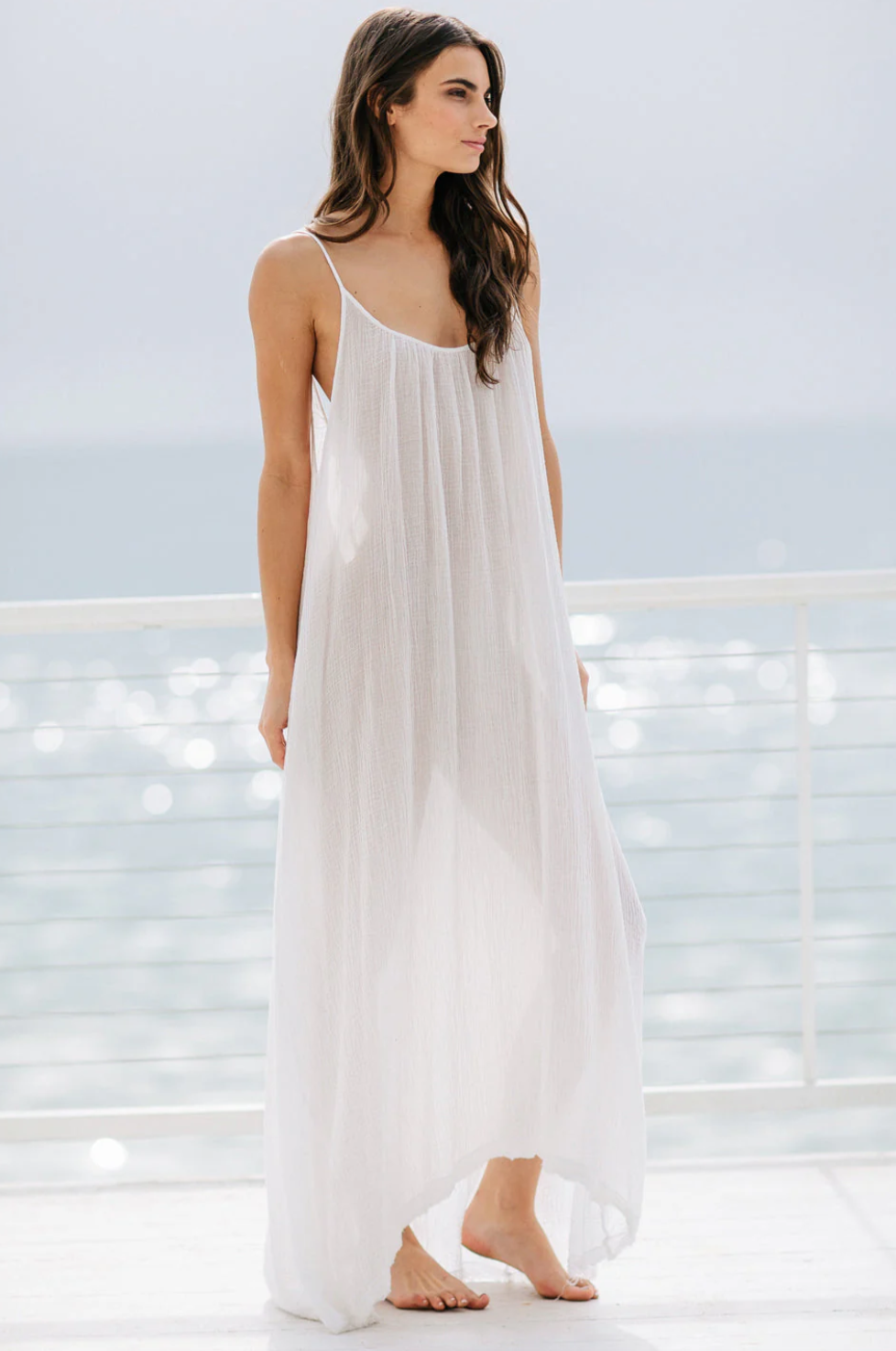 Barefoot on a sunlit deck with an ocean view, a woman wears a 9seed Tulum maxi dress made of lightweight gauze. Behind her, the sky is hazy and the water glistens.