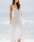 Barefoot on a sunlit deck with an ocean view, a woman wears a 9seed Tulum maxi dress made of lightweight gauze. Behind her, the sky is hazy and the water glistens.