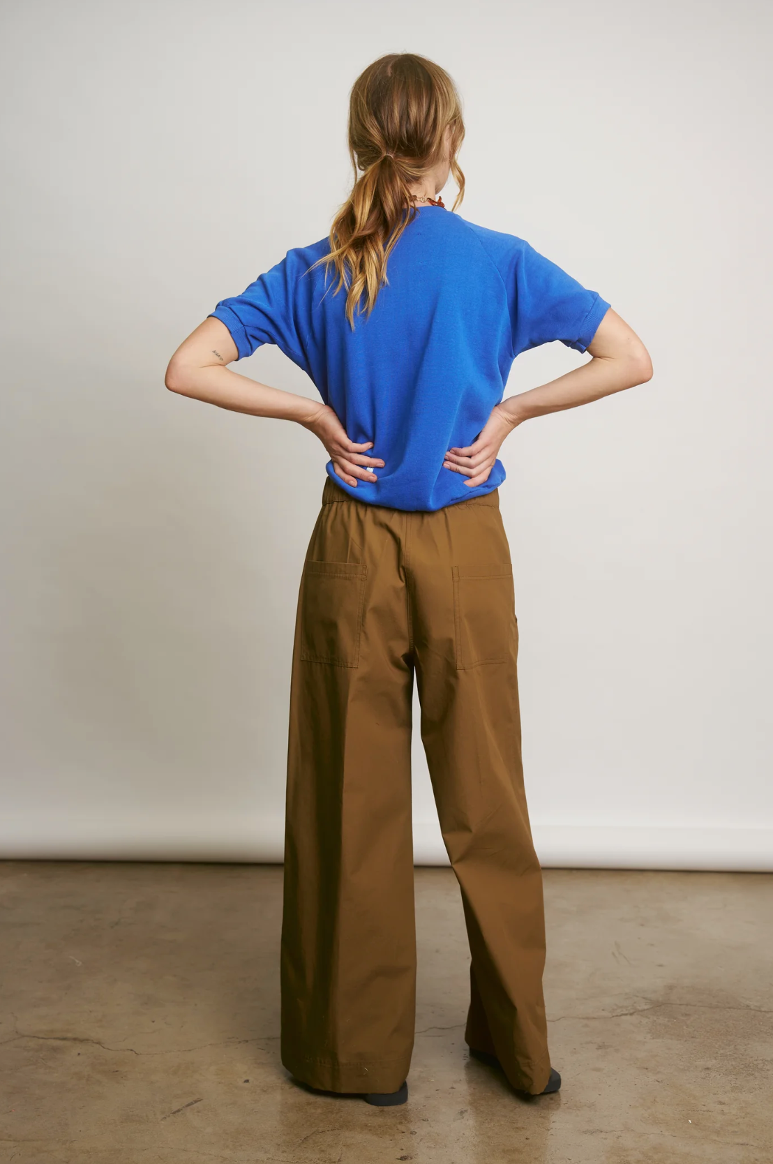 A person with long hair stands confidently, hands on hips, wearing a blue t-shirt and Connor Wide Leg Pant by Aquarius Cocktail. The backdrop is plain and neutral, echoing DTLA&#39;s minimalist style.