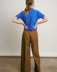 A person with long hair stands confidently, hands on hips, wearing a blue t-shirt and Connor Wide Leg Pant by Aquarius Cocktail. The backdrop is plain and neutral, echoing DTLA's minimalist style.