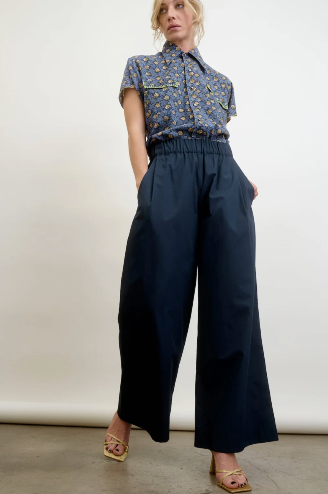 In a studio, someone wears Aquarius Cocktail&#39;s chic ensemble: a blue patterned short-sleeve shirt and Connor Wide Leg Pant in navy, made from a wool blend. With upstyled light hair and open-toed heels, the look showcases small batch quality against a plain white background.