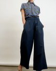 In a studio, someone wears Aquarius Cocktail's chic ensemble: a blue patterned short-sleeve shirt and Connor Wide Leg Pant in navy, made from a wool blend. With upstyled light hair and open-toed heels, the look showcases small batch quality against a plain white background.