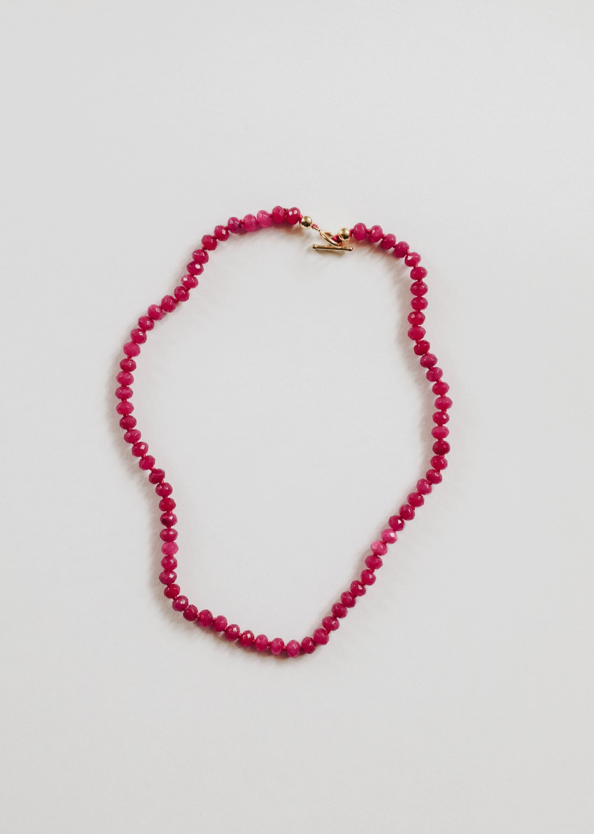 The Faire Gemstone Necklace showcases elegance with its small round pink beads and a gold-filled clasp, arranged in an oval shape on a white background, highlighting the beauty of handmade jewelry.