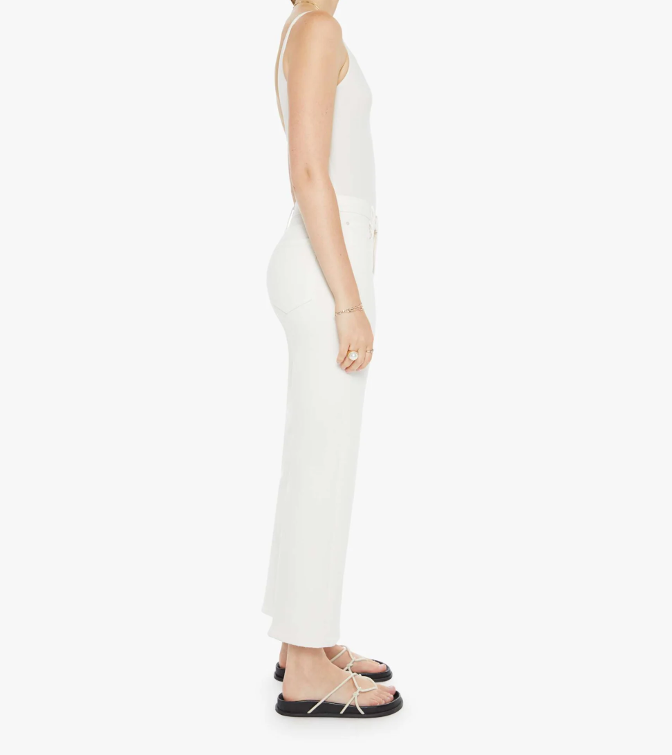 Someone wearing The Rambler Zip Ankle by Mother stands in profile against a plain backdrop, in a sleeveless white top and high-waisted white pants. Their outfit's stretch fabric is highlighted as they wear black sandals with white straps and a bracelet on their left wrist.