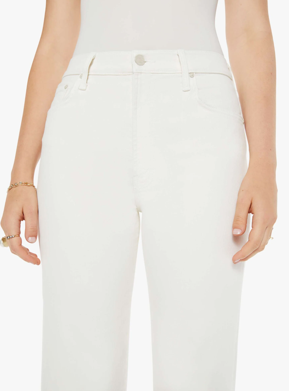 A person is wearing The Rambler Zip Ankle pants by Mother in an off-white wash paired with a matching white top. Gold bracelets and rings adorn their relaxed arms. The stretch fabric enhances comfort against a plain white backdrop.