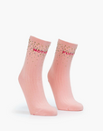 Two baby pink tube socks from Mother, named "The Half Step," stand upright against a white background, featuring a ribbed texture. They are adorned with colorful crystal embellishments at the top and have bold red text across them.