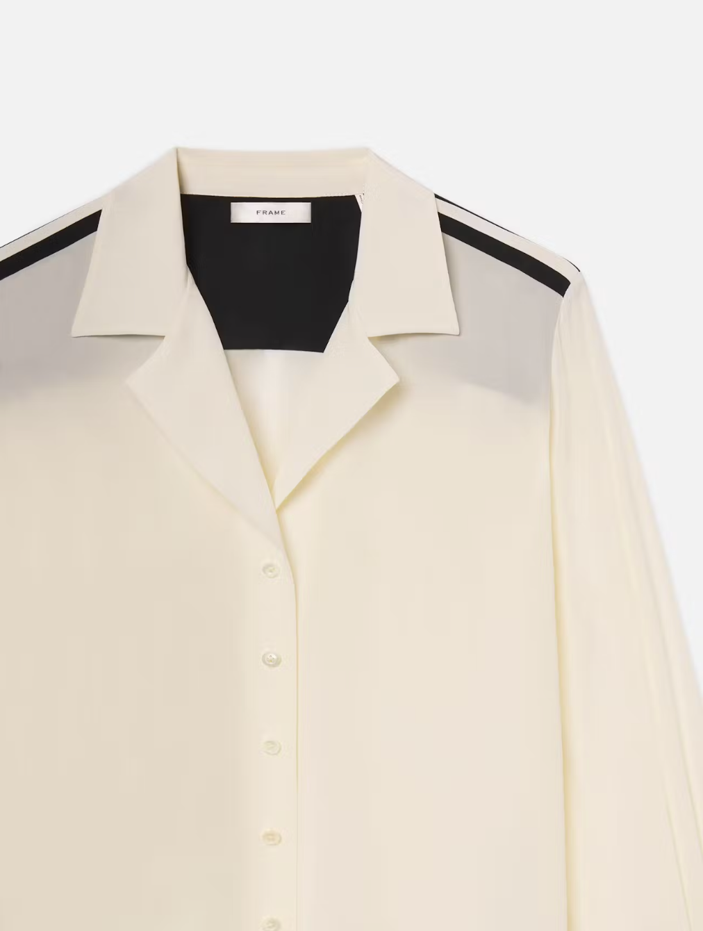 FRAME's Silk Pajama Shirt in cream features black shoulder stripes and a classic collar. Its minimalist design enhances versatility, making it an effortless wardrobe staple. Presented elegantly against a plain white background.