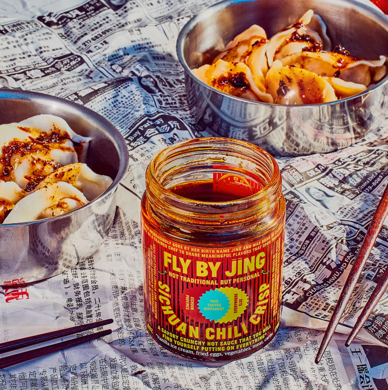 A jar of Faire's Fly By Jing Sichuan Chili Crisp is placed on a newspaper backdrop. Two dumpling bowls with savory sauce sit next to it, chopsticks resting close by, accentuating the vibrant scene centered around this star chili product.