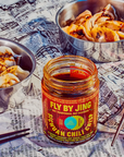 A jar of Faire's Fly By Jing Sichuan Chili Crisp is placed on a newspaper backdrop. Two dumpling bowls with savory sauce sit next to it, chopsticks resting close by, accentuating the vibrant scene centered around this star chili product.