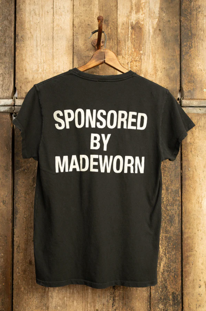 A MADE WORN "ARTIST" CROP POCKET TEE, black in color, hangs on a wooden wall displaying "SPONSORED BY MADEWORN" in white capitals. Its vintage unisex fit and unique distressing highlight its handmade quality crafted in Los Angeles.