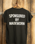 A MADE WORN "ARTIST" CROP POCKET TEE, black in color, hangs on a wooden wall displaying "SPONSORED BY MADEWORN" in white capitals. Its vintage unisex fit and unique distressing highlight its handmade quality crafted in Los Angeles.