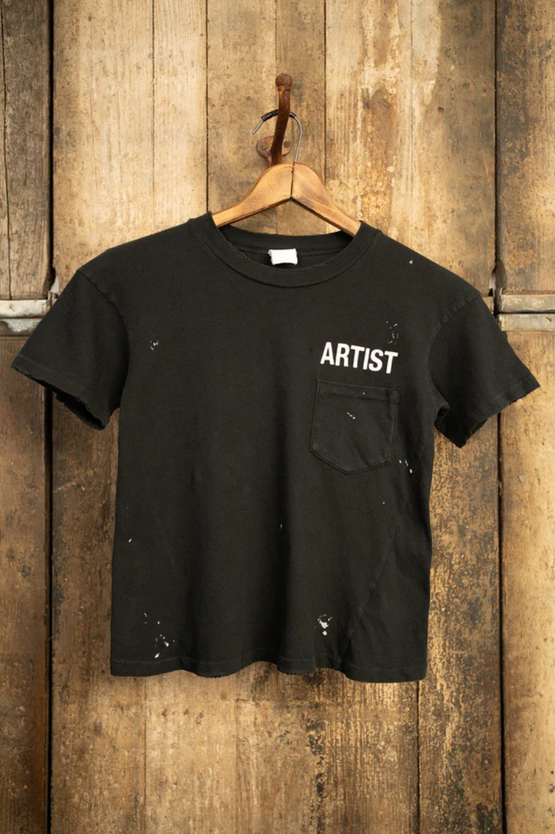 The MADE WORN "ARTIST" CROP POCKET TEE by Made Worn is a black, vintage unisex fit handmade T-shirt from Los Angeles. It features "ARTIST" in white near the left shoulder, with unique white paint stains and a small chest pocket, displayed on a wooden hanger against a textured wood backdrop.