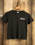 The MADE WORN "ARTIST" CROP POCKET TEE by Made Worn is a black, vintage unisex fit handmade T-shirt from Los Angeles. It features "ARTIST" in white near the left shoulder, with unique white paint stains and a small chest pocket, displayed on a wooden hanger against a textured wood backdrop.