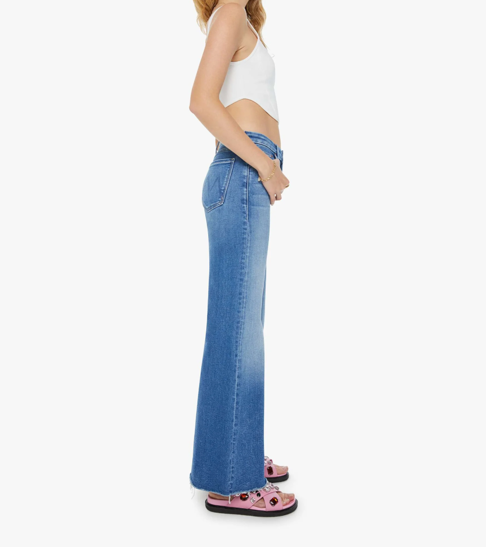 A side view shows someone with shoulder-length hair against a plain background, wearing The Roller Fray by Mother—white sleeveless top, high-waisted mid-blue wide-leg jeans—and paired with pink sandals, standing relaxed and looking forward.