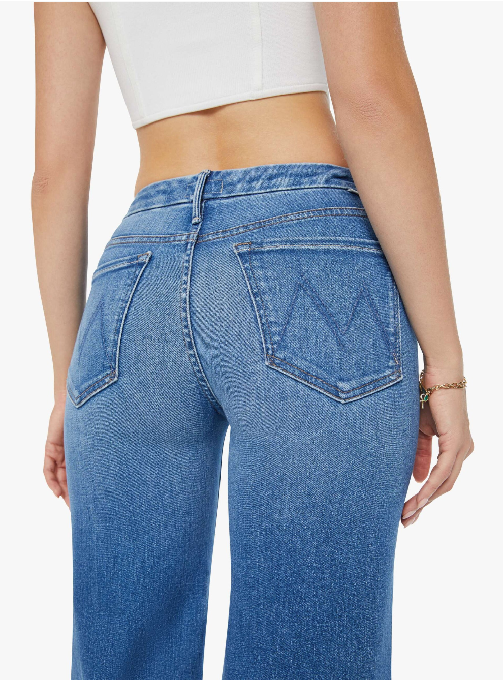 Rear view of someone wearing Mother’s The Roller Fray high-waisted wide leg blue jeans with embroidered back pockets and a white crop top. The mid-blue wash denim pairs perfectly with the bracelet on the right wrist, adding flair to the look.