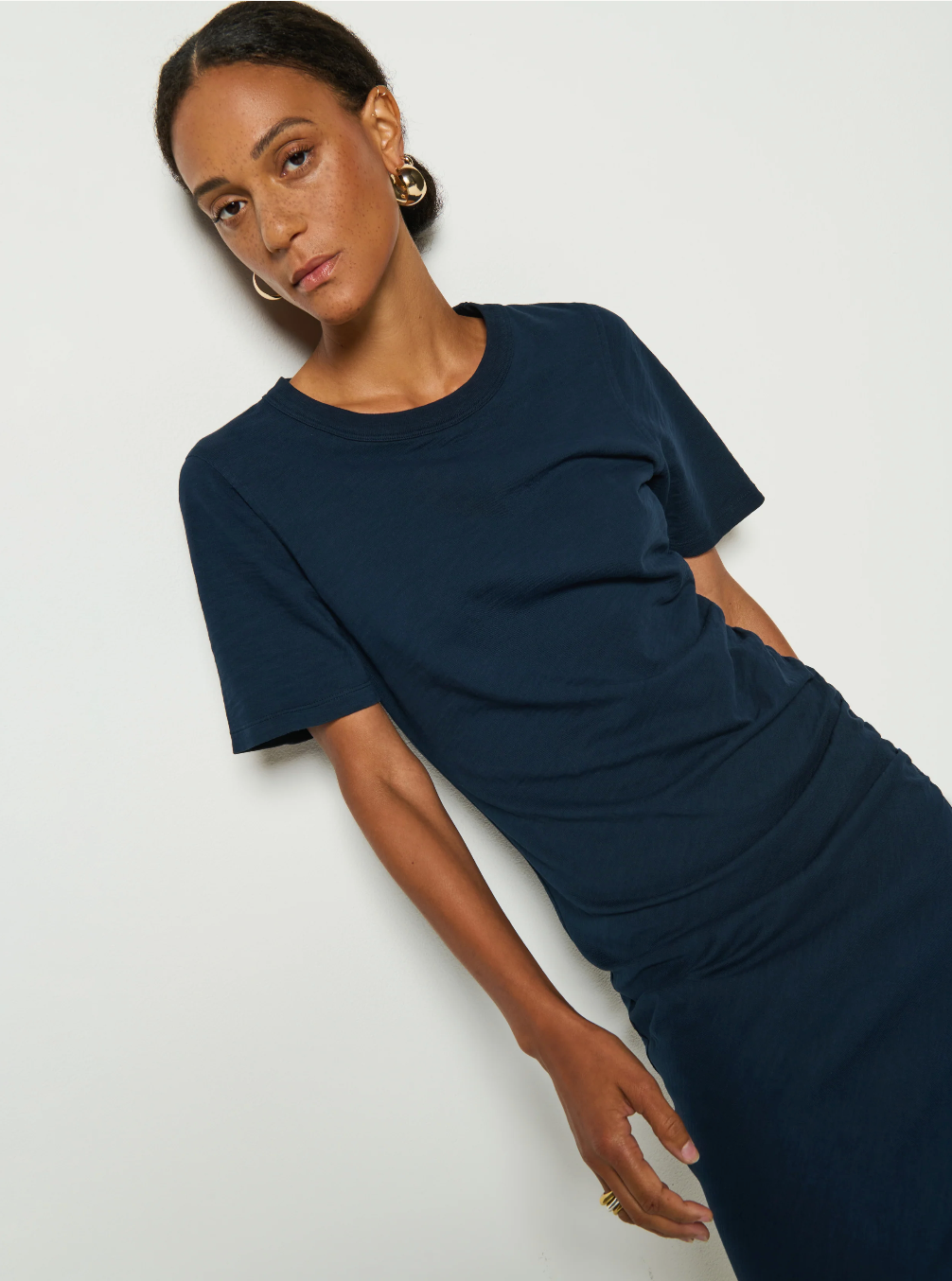 A person models the dark blue Cole T-shirt Dress by Nation LTD, made from hypoallergenic Peruvian Cotton, against a plain white background. Their hoop earrings add flair as they look at the camera with a neutral expression.