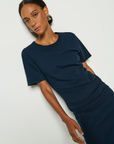 A person models the dark blue Cole T-shirt Dress by Nation LTD, made from hypoallergenic Peruvian Cotton, against a plain white background. Their hoop earrings add flair as they look at the camera with a neutral expression.