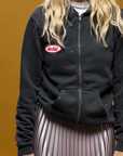 A person with long blonde hair is wearing a MadeWorn Artist Hoodie by Made Worn, handcrafted in Los Angeles. It's paired with a metallic pleated skirt against a solid mustard yellow backdrop, while the person's face stays hidden.