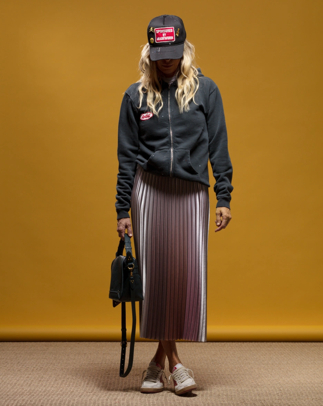 A person with long, wavy hair wears a MadeWorn Sponsor Hat from Made Worn with an adjustable back strap, a dark zippered hoodie, and a pleated skirt. They hold a small black handbag and wear white sneakers against a mustard yellow background.