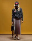 A person with long, wavy hair wears a MadeWorn Sponsor Hat from Made Worn with an adjustable back strap, a dark zippered hoodie, and a pleated skirt. They hold a small black handbag and wear white sneakers against a mustard yellow background.